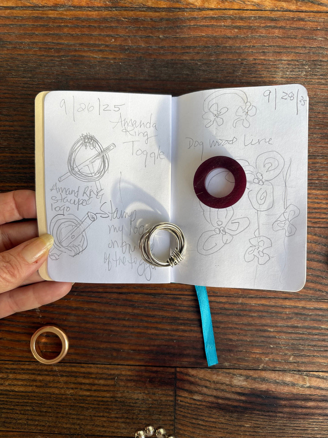 Open sketchbook showing hand-drawn jewelry designs labeled “Dogwood Line” with a silver ring and red wax sample placed on the pages.