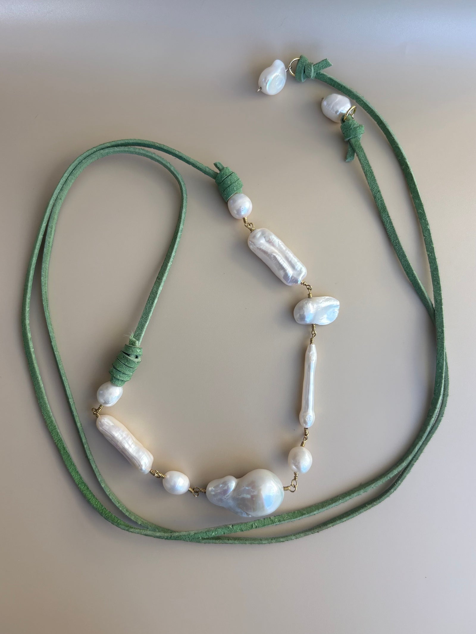 A close-up of a green deerskin leather wrap necklace adorned with freshwater and baroque pearls, shown on beige background