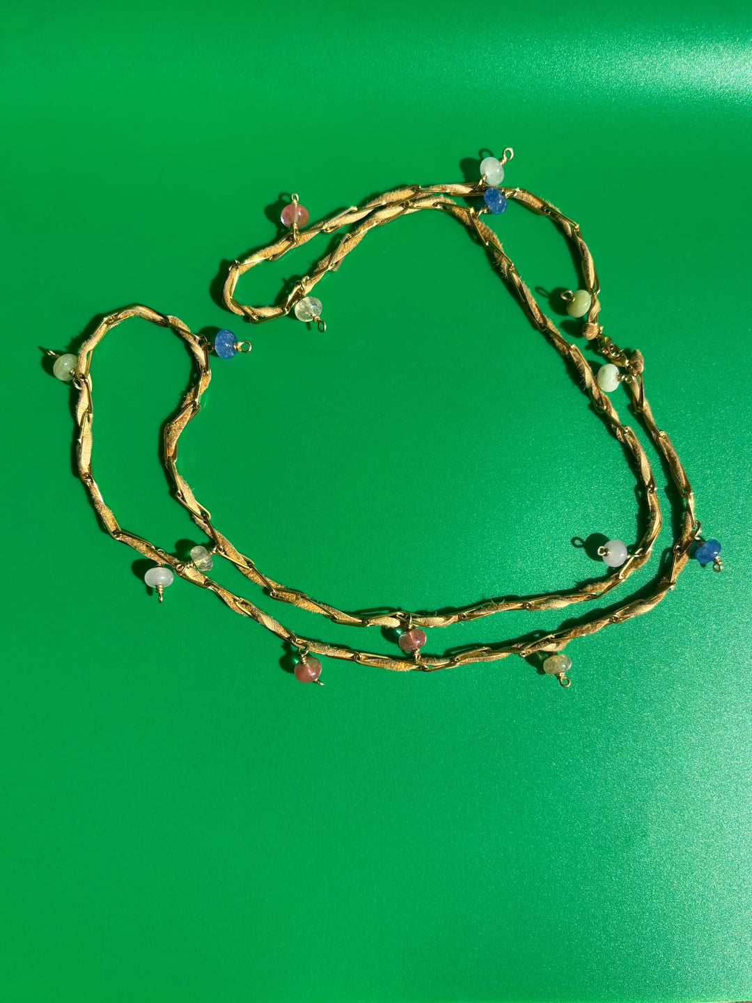 Tan deerskin leather and gold-toned paperclip belly chain laid flat on green background. Survivor-made ethical jewelry symbolizing endurance.