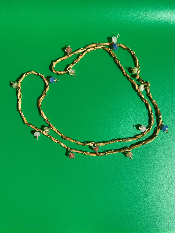 Tan deerskin leather and gold-toned paperclip belly chain laid flat on green background. Survivor-made ethical jewelry symbolizing endurance.
