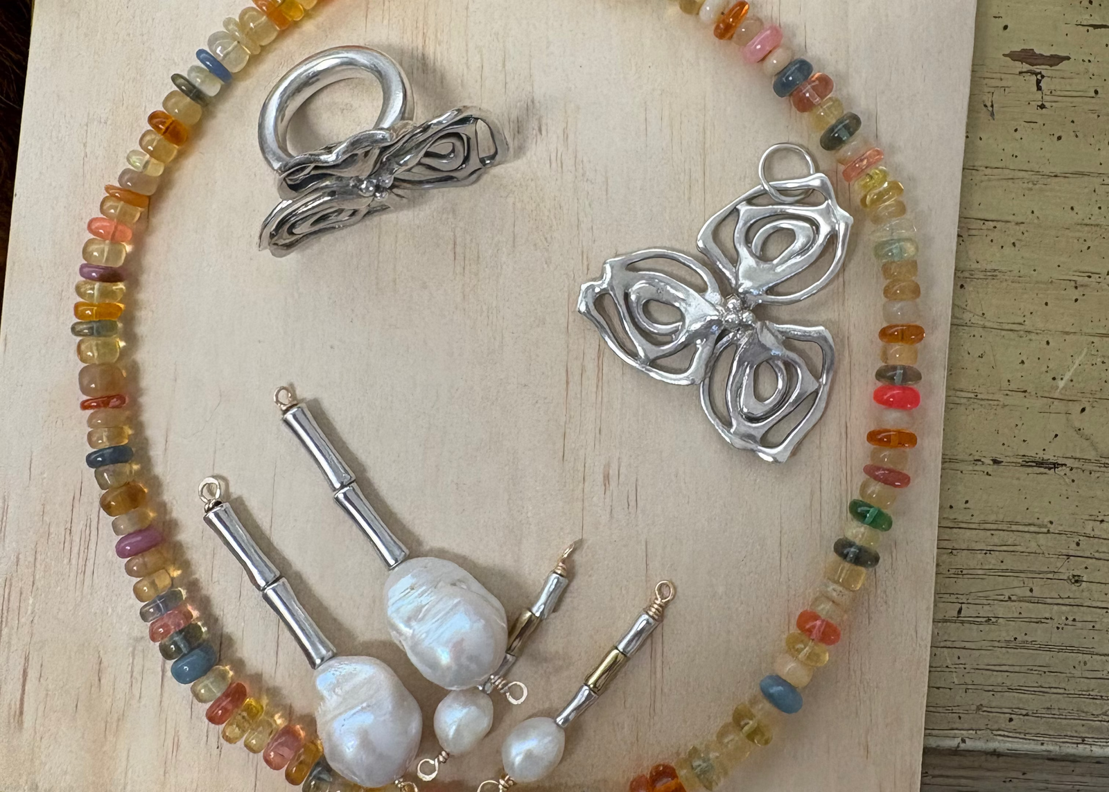 Jewelry One of a KIND display featuring the Dogwood Ring and Charm, Vertebrae Mini and Maxi earrings, and Smartie Opal Necklace arranged on a light wooden board.