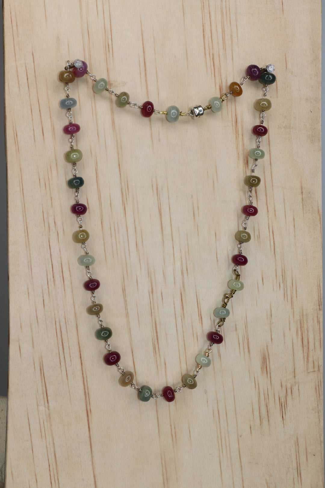 Sapphire rondelle necklace featuring multicolored beads in burgundy, green, blue, and golden tones, linked with metal wire and photographed on a light wood background.