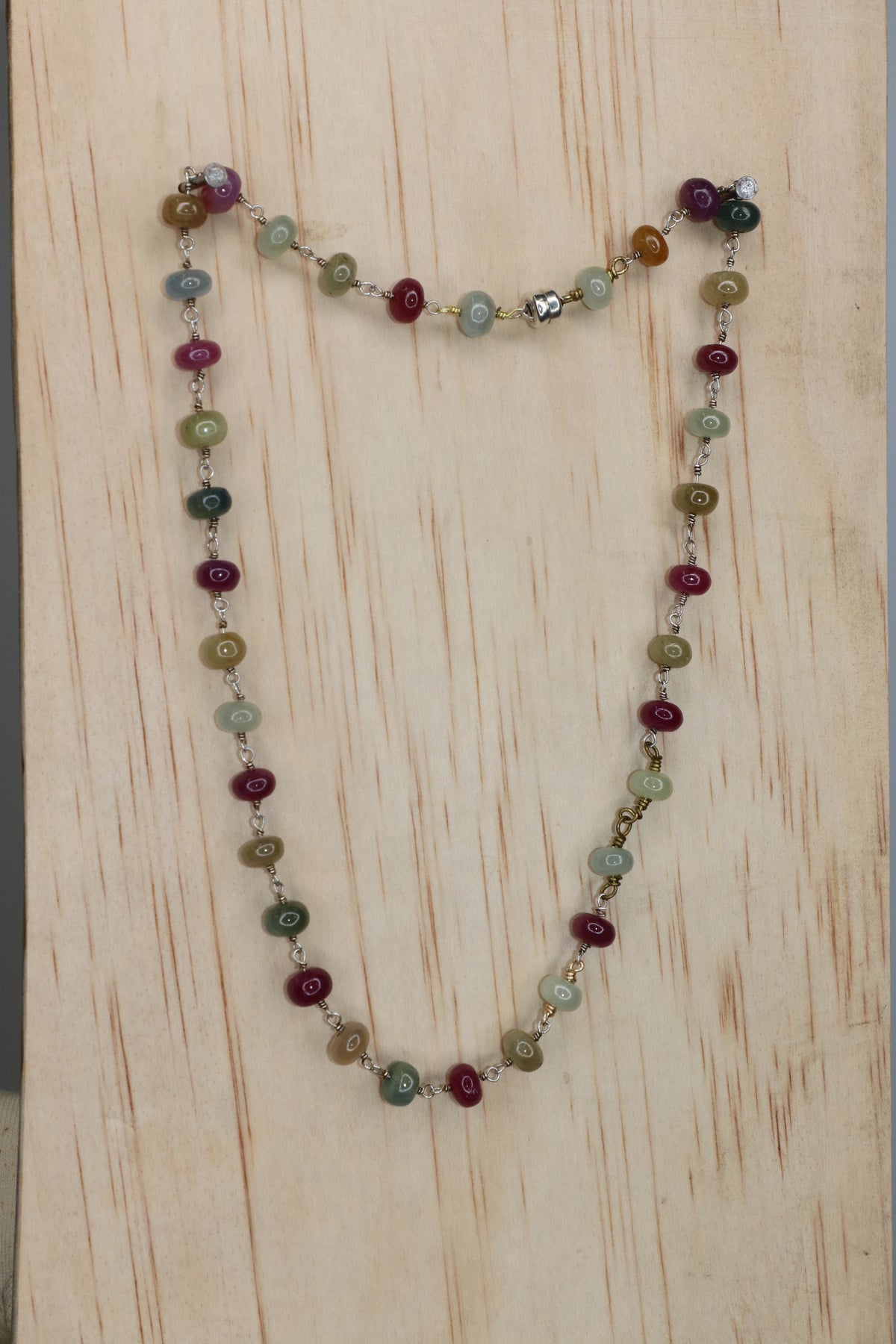 Sapphire rondelle necklace featuring multicolored beads in burgundy, green, blue, and golden tones, linked with metal wire and photographed on a light wood background.