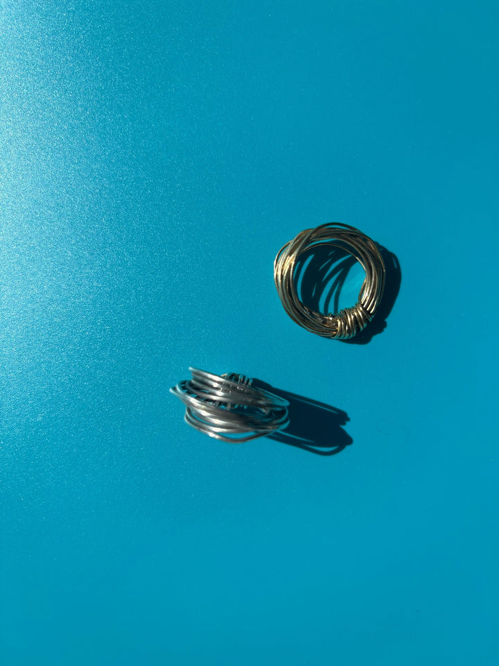 Brass and Fairmined sterling silver knot rings hand-wrapped from 18-gauge wire, displayed together on a blue background. Survivor-made ethical jewelry by Jewelry One of a KIND symbolizing strength and resilience.