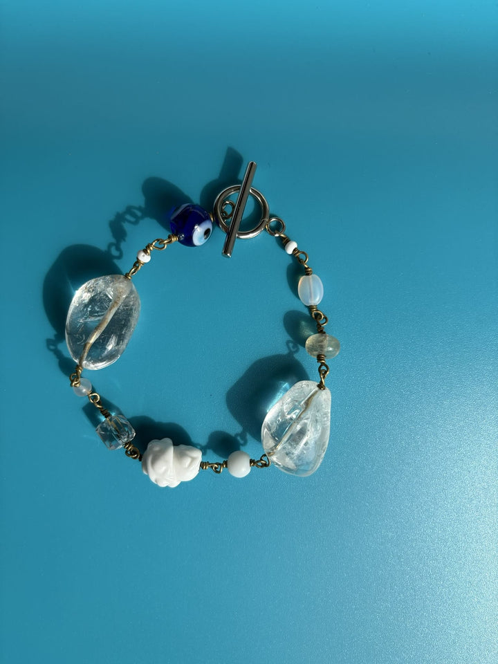 Rock crystal bracelet with Evil Eye charm shown on blue background. Survivor-made luxury jewelry representing protection.