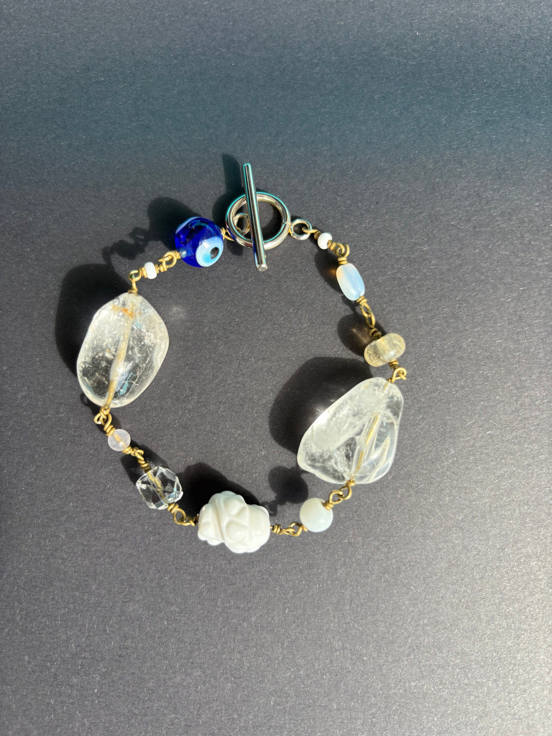 Rock crystal with blue evil eye and white coral bracelet on black surface. Survivor-made ethical jewelry symbolizing clarity and empowerment.
