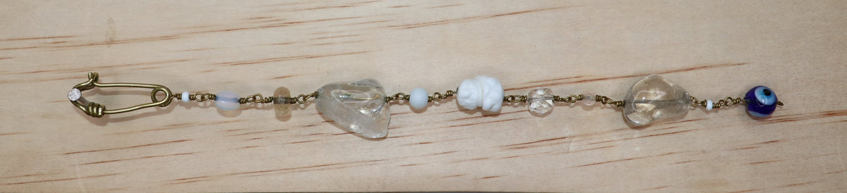 A handmade beaded  Jamila bracelet featuring clear quartz, white beads, a blue evil eye bead, and a gold-toned safety pin clasp, hanging on a light wood board.