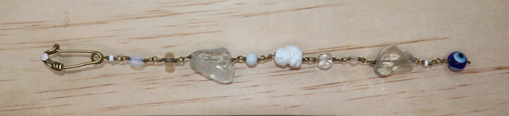 A handmade beaded  Jamila bracelet featuring clear quartz, white beads, a blue evil eye bead, and a gold-toned safety pin clasp, hanging on a light wood board.