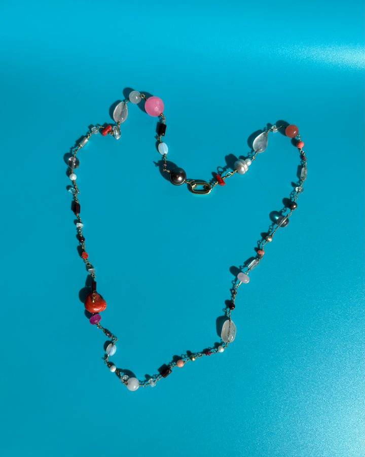 Pink gemstone necklace with ruby, pearl, Swarovski crystals, and coral on blue surface. Ethical jewelry symbolizing confidence and joy.