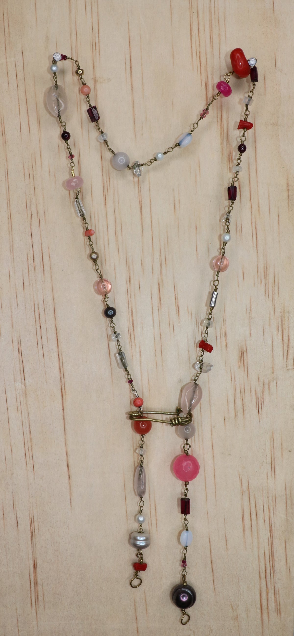 A long beaded necklace with Rose quartz, pearl, pink dyed quartz, and coral necklace displayed  on a light wood block 