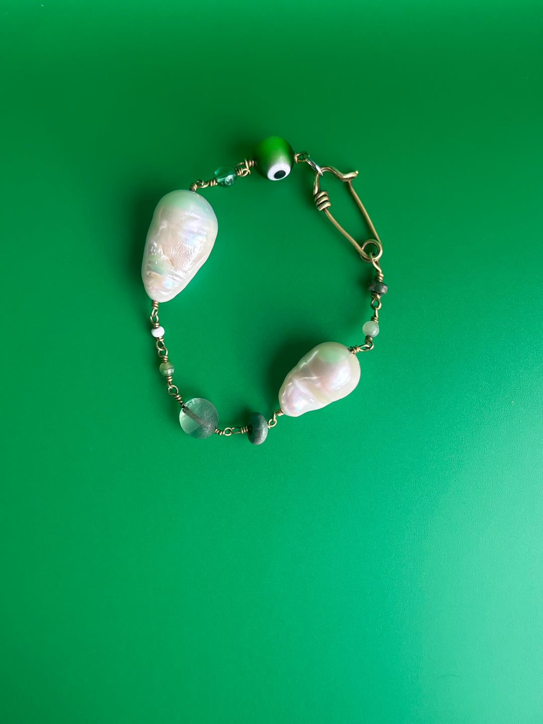 Baroque Pearl bracelet with green Evil Eye charm and gemstones, turquoise and peridot, on beige surface. Survivor-made ethical jewelry of strength and resilience.