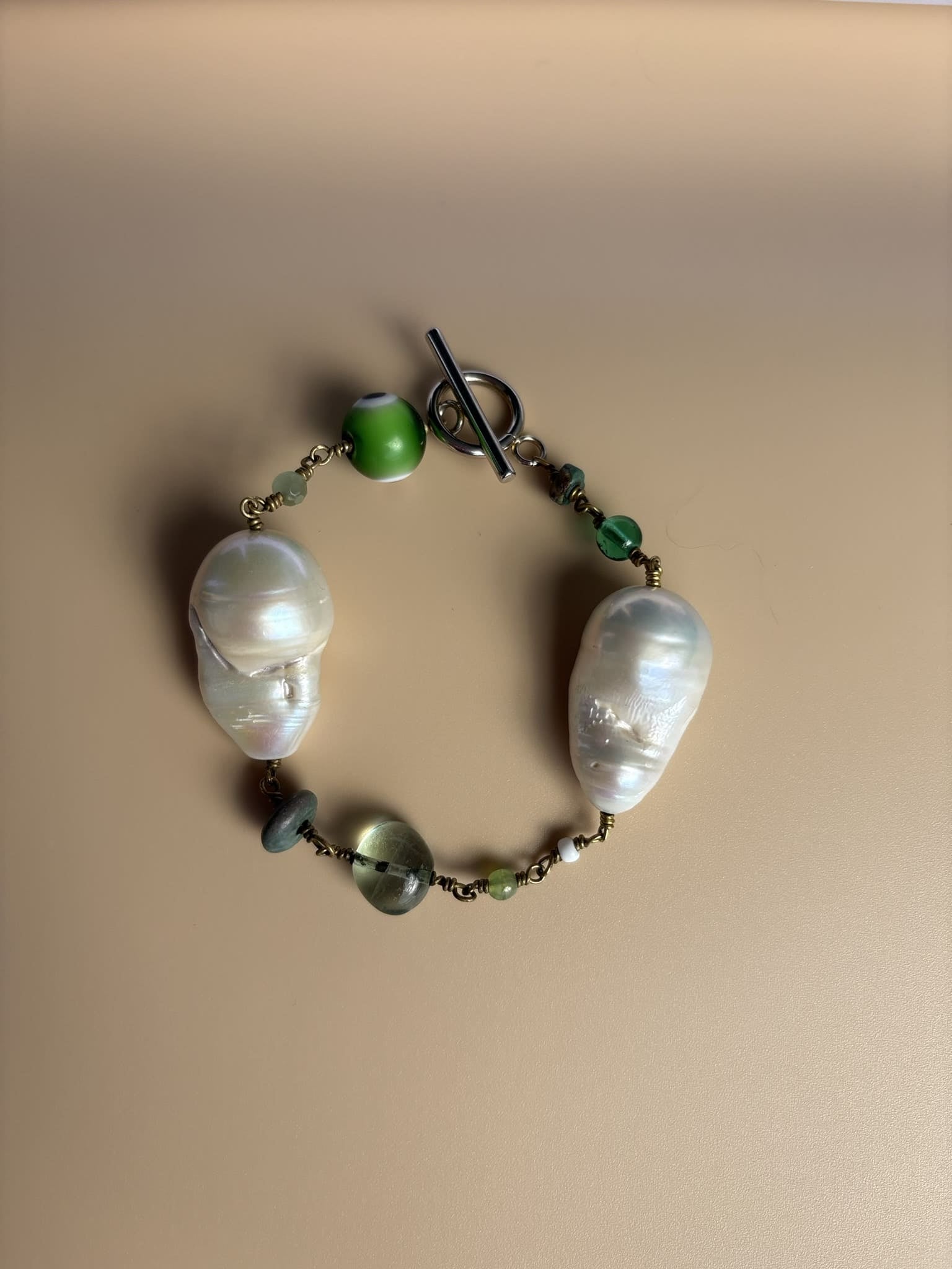 Baroque Pearl bracelet with green Evil Eye charm and gemstones, turquoise and peridot, on beige surface. Survivor-made ethical jewelry of strength and resilience.
