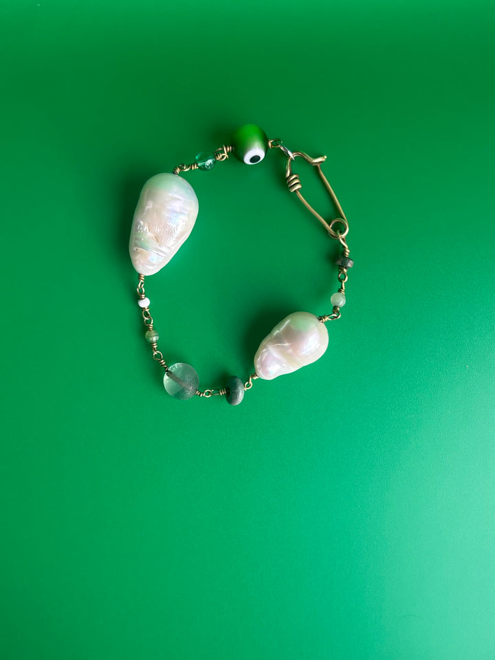 Baroque Pearl bracelet with green Evil Eye charm and gemstones, turquoise and peridot, on beige surface. Survivor-made ethical jewelry of strength and resilience.