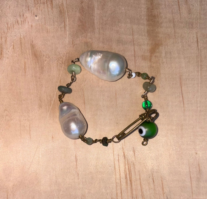 Marya collectible bracelet with two freshwater baroque pearls, individually selected green gemstones, Evil Eye charm, and handcrafted KIND Closure made out of brass. 