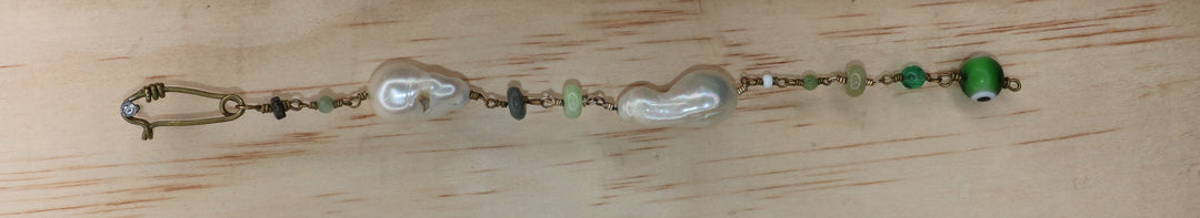 Marya collectible bracelet with two freshwater baroque pearls, individually selected green gemstones, Evil Eye charm, and handcrafted KIND Closure made out of brass laid flat on a piec of wood. 