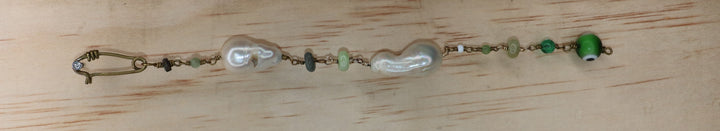 Marya collectible bracelet with two freshwater baroque pearls, individually selected green gemstones, Evil Eye charm, and handcrafted KIND Closure made out of brass laid flat on a piec of wood. 
