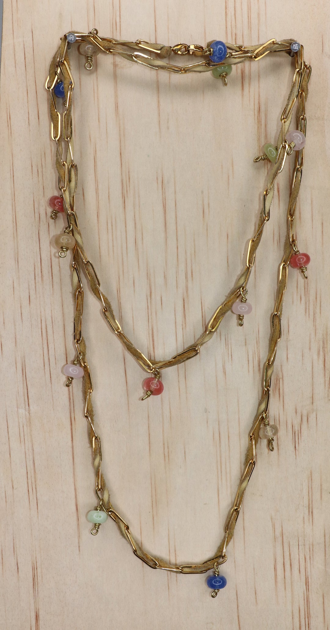 Nadia belly chain styled as a layered necklace, featuring tan deerskin leather, sculptural gold-toned paperclip links, and dangling multicolored beads in blue, pink, green, and coral, displayed on a light wood background.