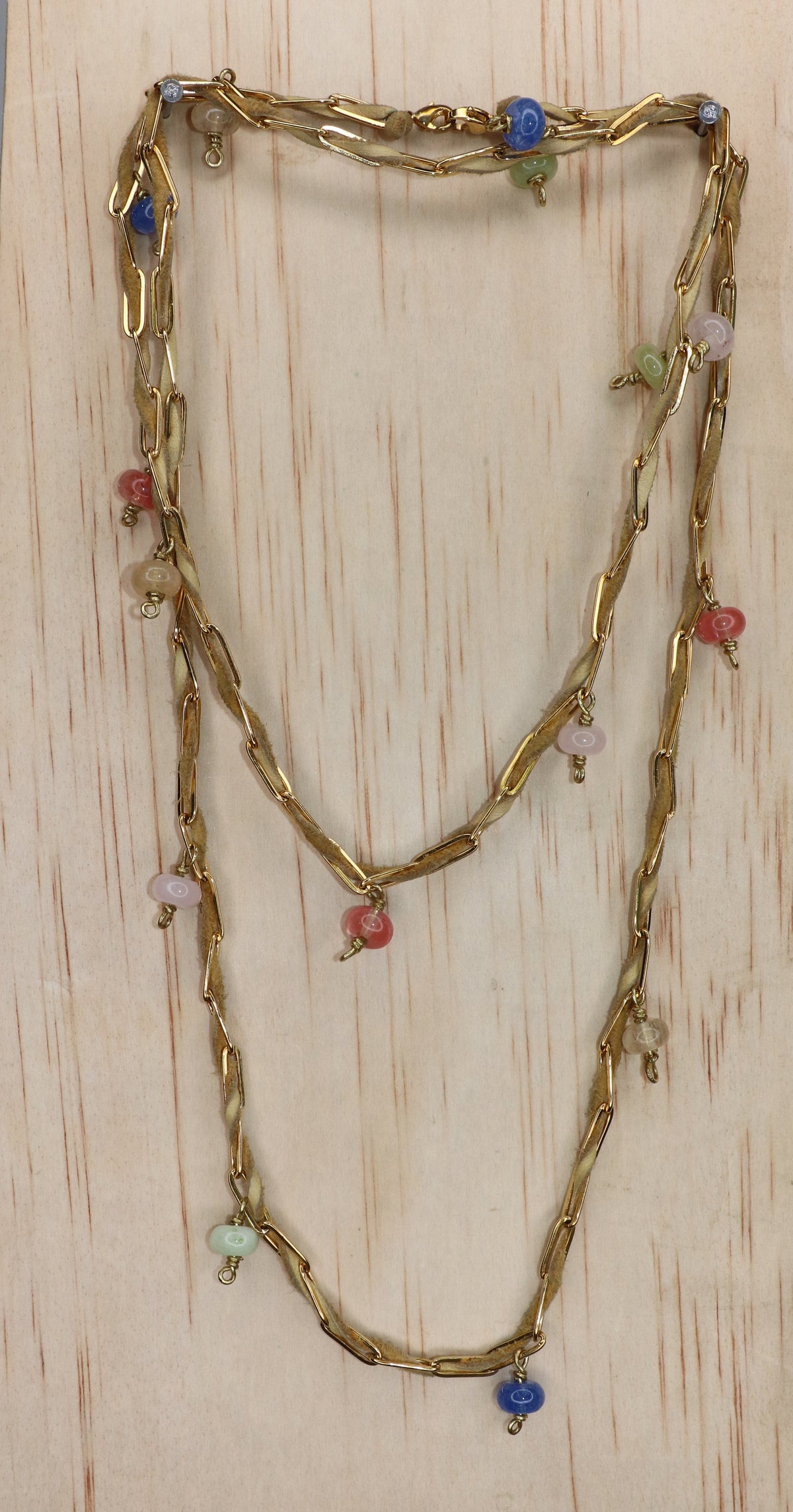 Nadia belly chain styled as a layered necklace, featuring tan deerskin leather, sculptural gold-toned paperclip links, and dangling multicolored beads in blue, pink, green, and coral, displayed on a light wood background.