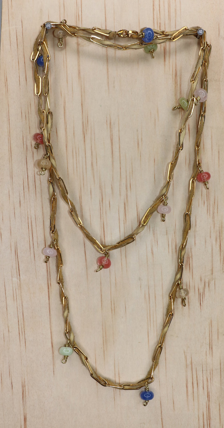 Nadia belly chain styled as a layered necklace, featuring tan deerskin leather, sculptural gold-toned paperclip links, and dangling multicolored beads in blue, pink, green, and coral, displayed on a light wood background.