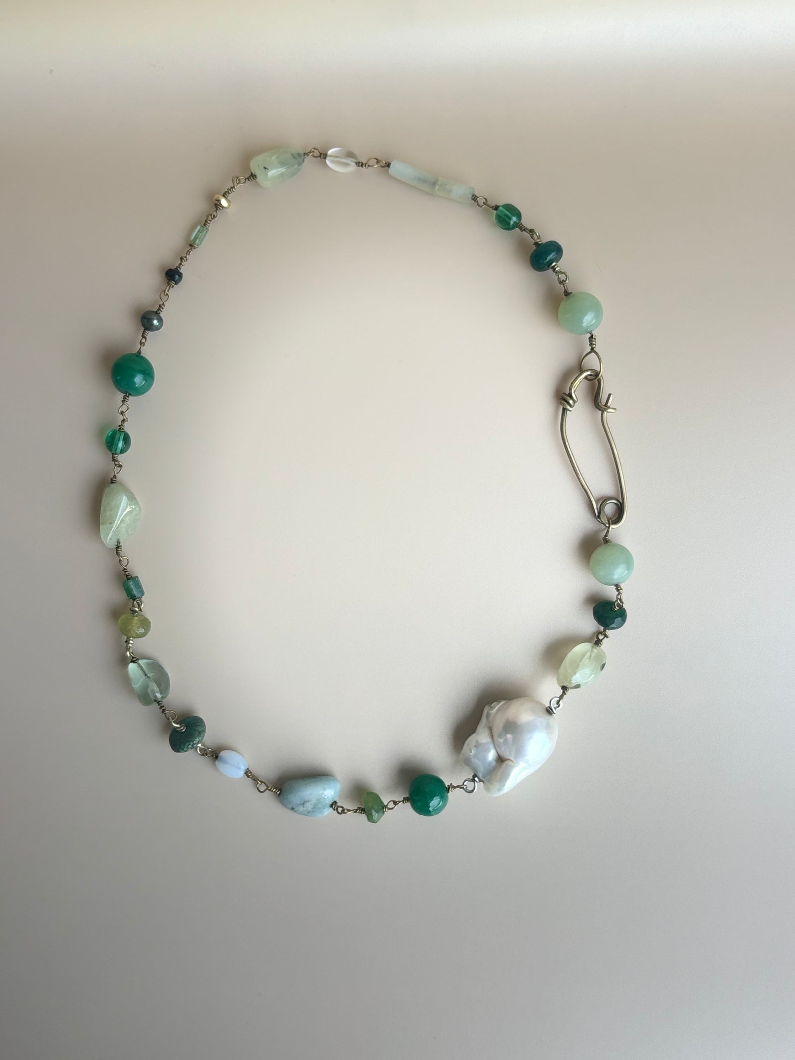 Green gemstone necklace with jadeite, peridot, turquoise, and a baroque pearl laid flat on a beige background. Survivor-made ethical jewelry by Jewelry One of a KIND symbolizing resilience and renewal.
