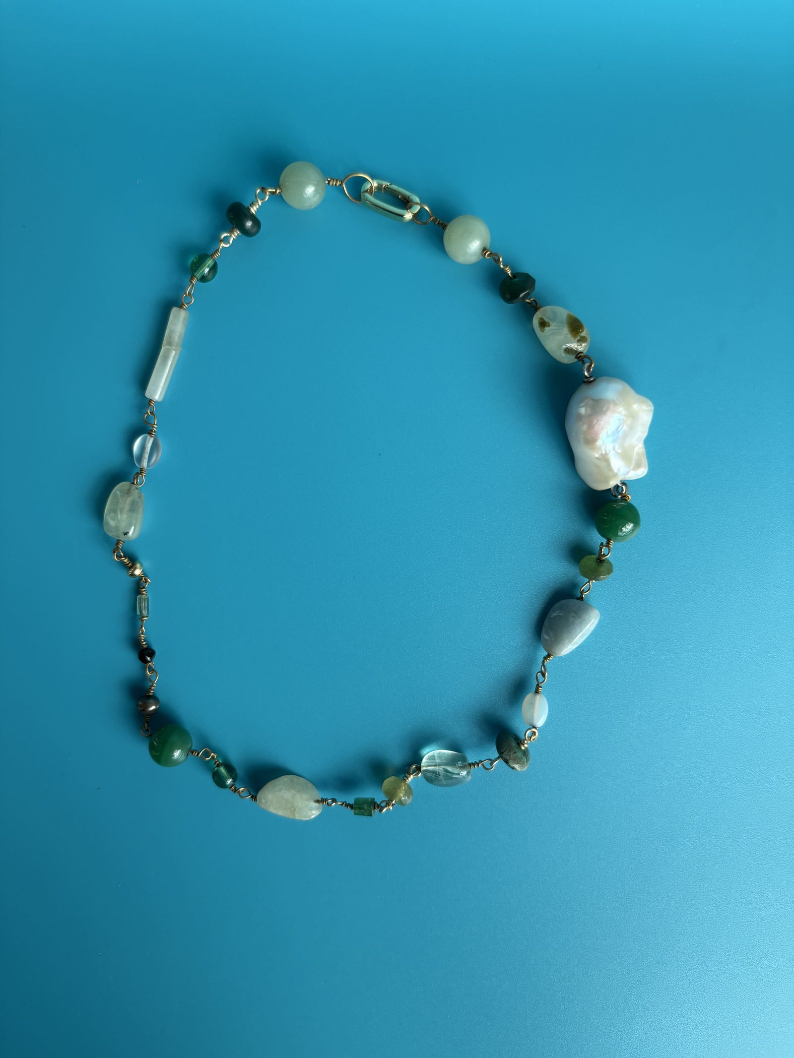 Green gemstone and baroque pearl necklace on blue surface. Ethical jewelry crafted by survivors celebrating renewal.